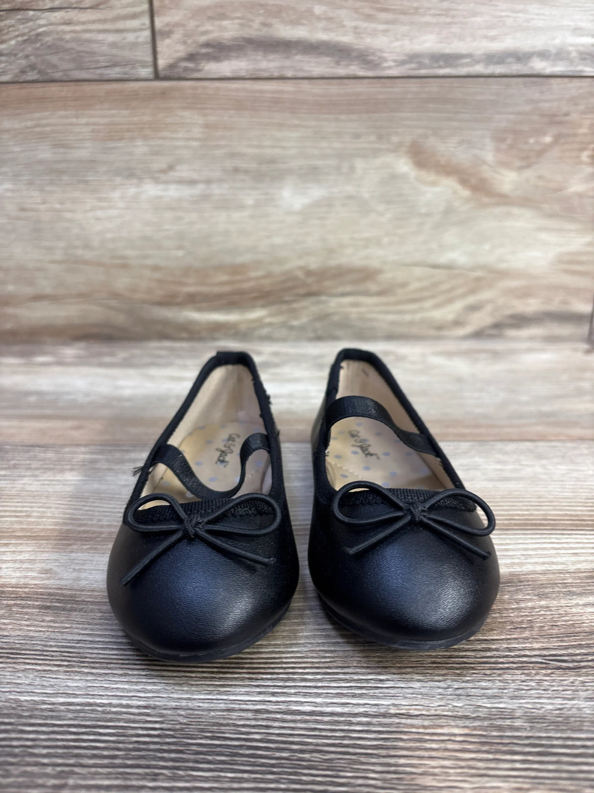Front view of Cat and Jack Nora black toddler slip-on ballet flats showing bow detail and elastic strap on wood floor background