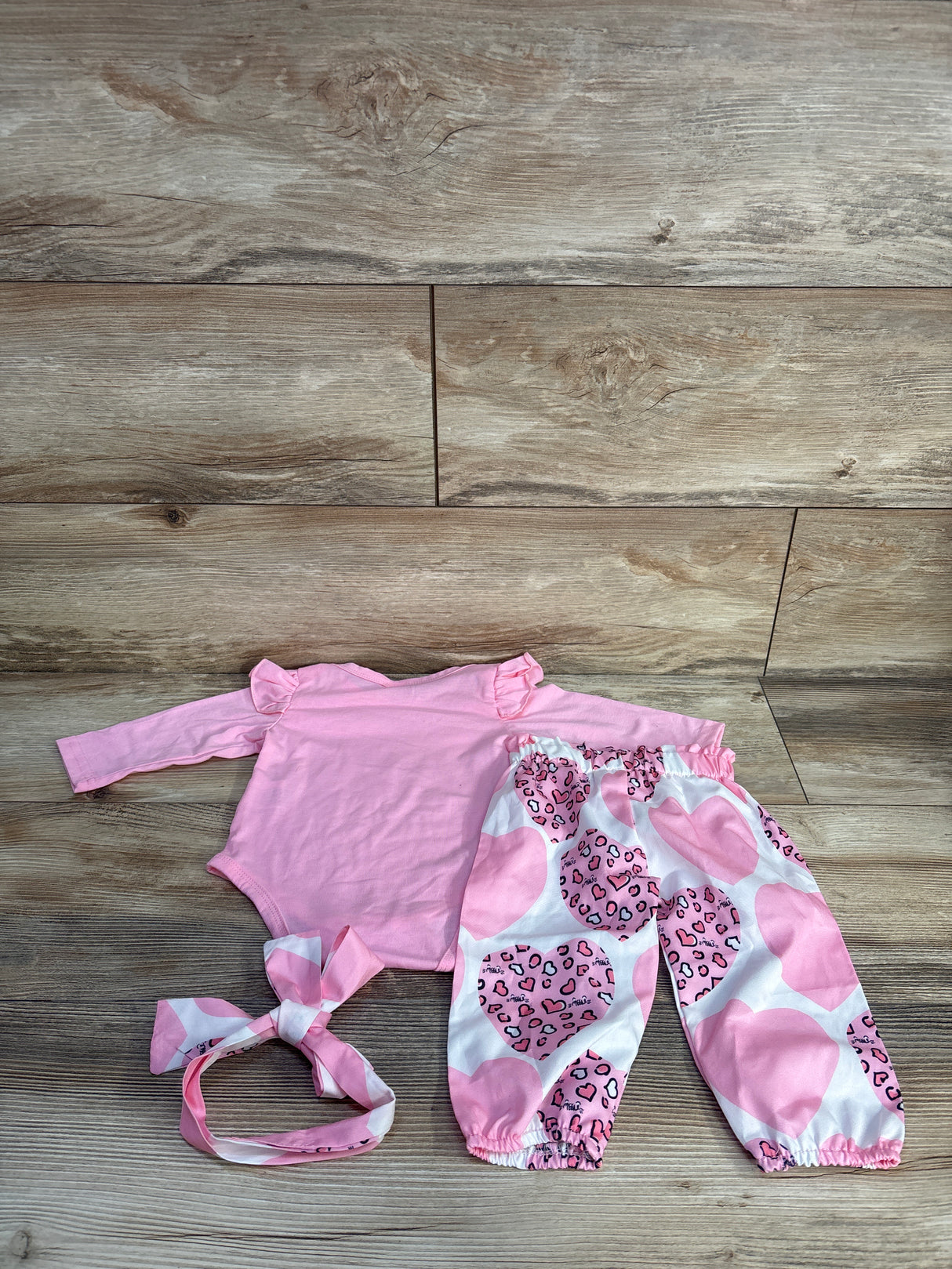 Flat lay of pink 3-piece baby set: ruffle-shoulder bodysuit, heart-print elastic pants, and tied headband on wood background