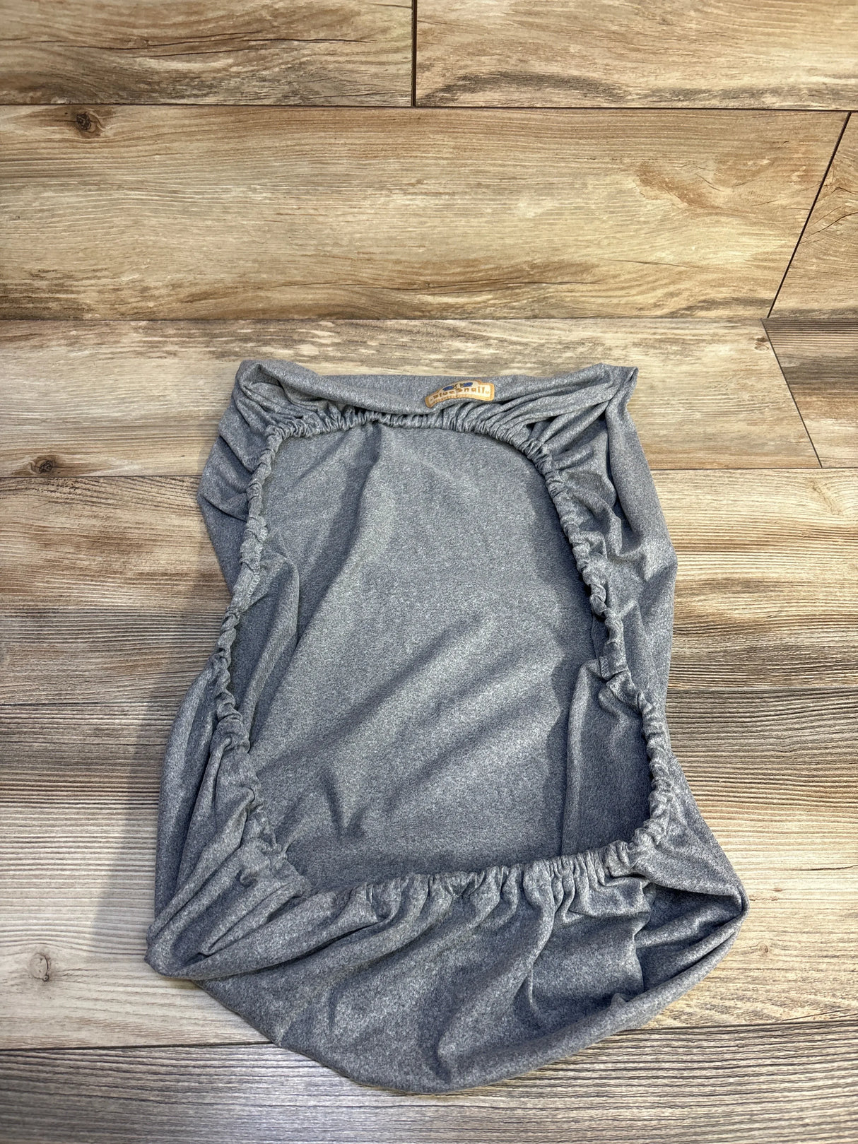 Top-down view of BlueSnail soft stretchy grey bassinet fitted sheet showing elasticized edges on wood floor