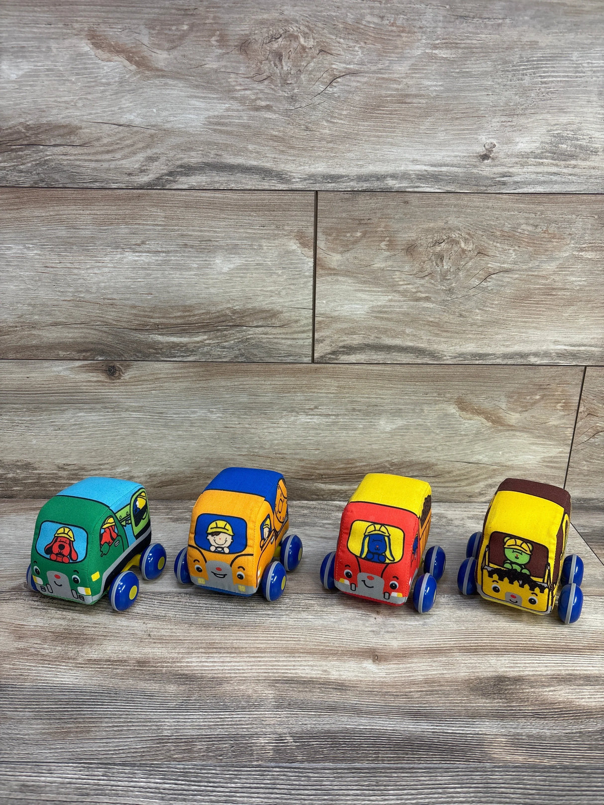 Front view of four K's Kids pull-back construction vehicles, colorful fabric bodies with blue wheels on wood plank background