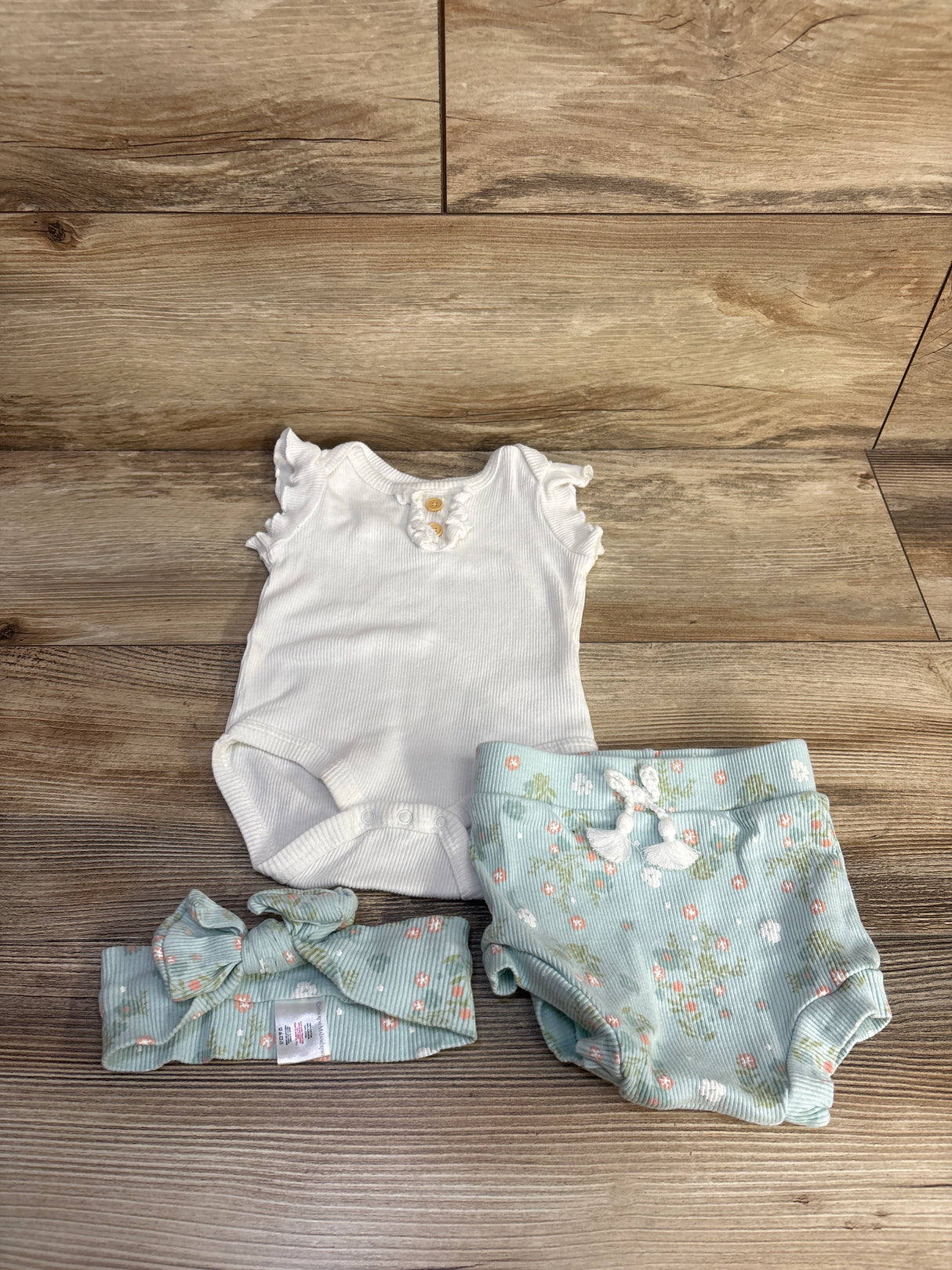 Top-down view of 3-piece ribbed baby set: white bodysuit, ruffled sleeves, mint floral bottoms and matching headband on wood