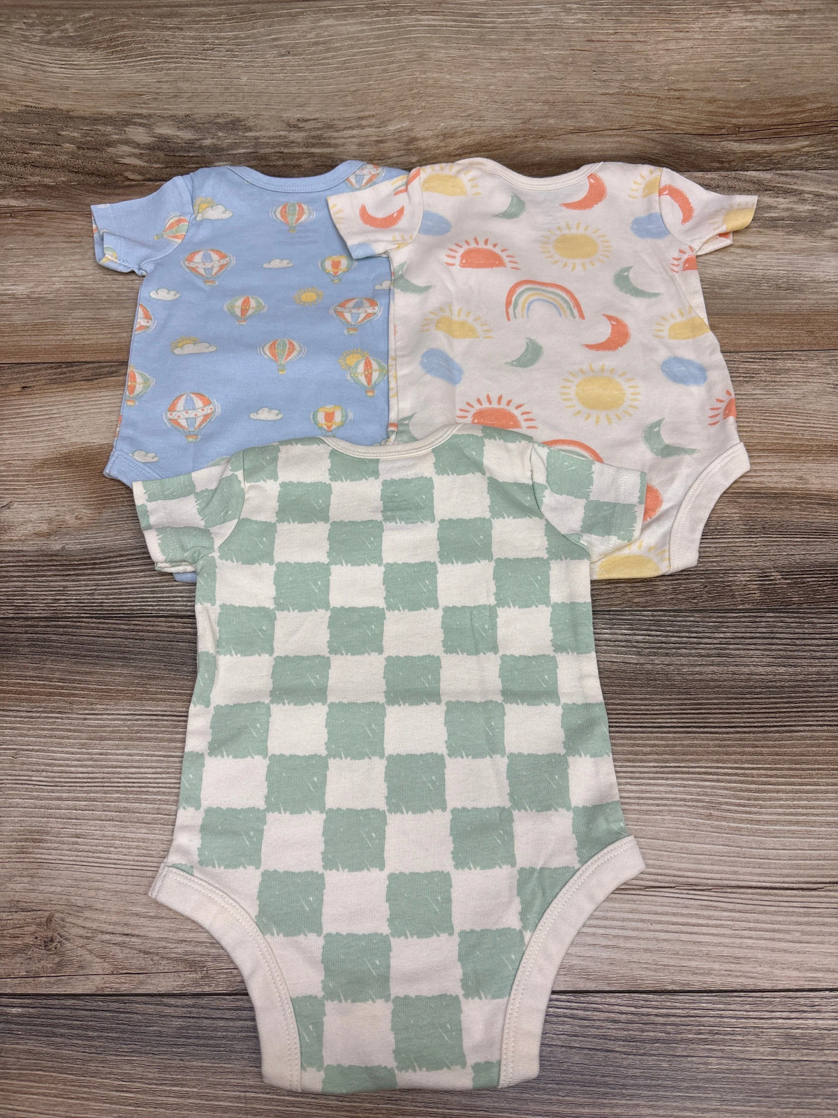 Top view of Member’s Mark 3pk organic bodysuits, size 3m, with balloon, celestial, and checkered patterns on wood