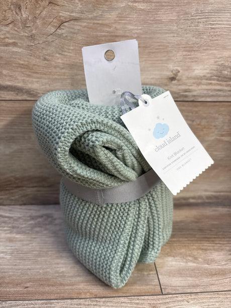 Angled view of Cloud Island knit baby blanket in sage green showing ribbed texture and brand tag on wood background
