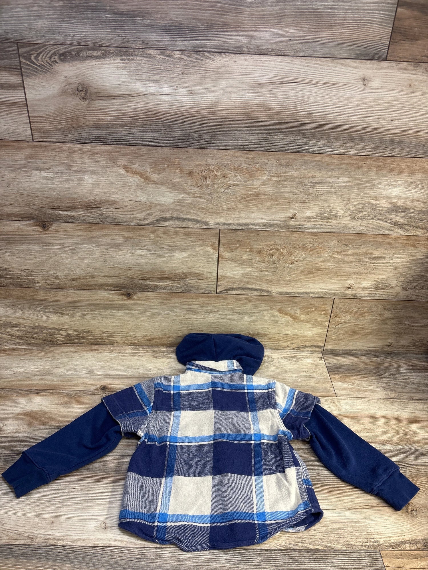 Back view of a blue plaid hooded flannel shacket with navy sleeves, laid flat on a wood floor