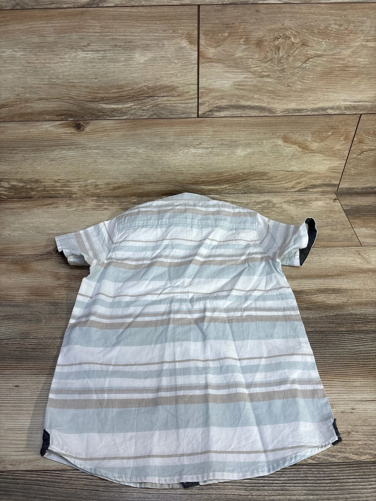 Back view of white boys' short-sleeve button-up shirt with gray and tan stripes, showing curved hem and cuff detail on wood floor background