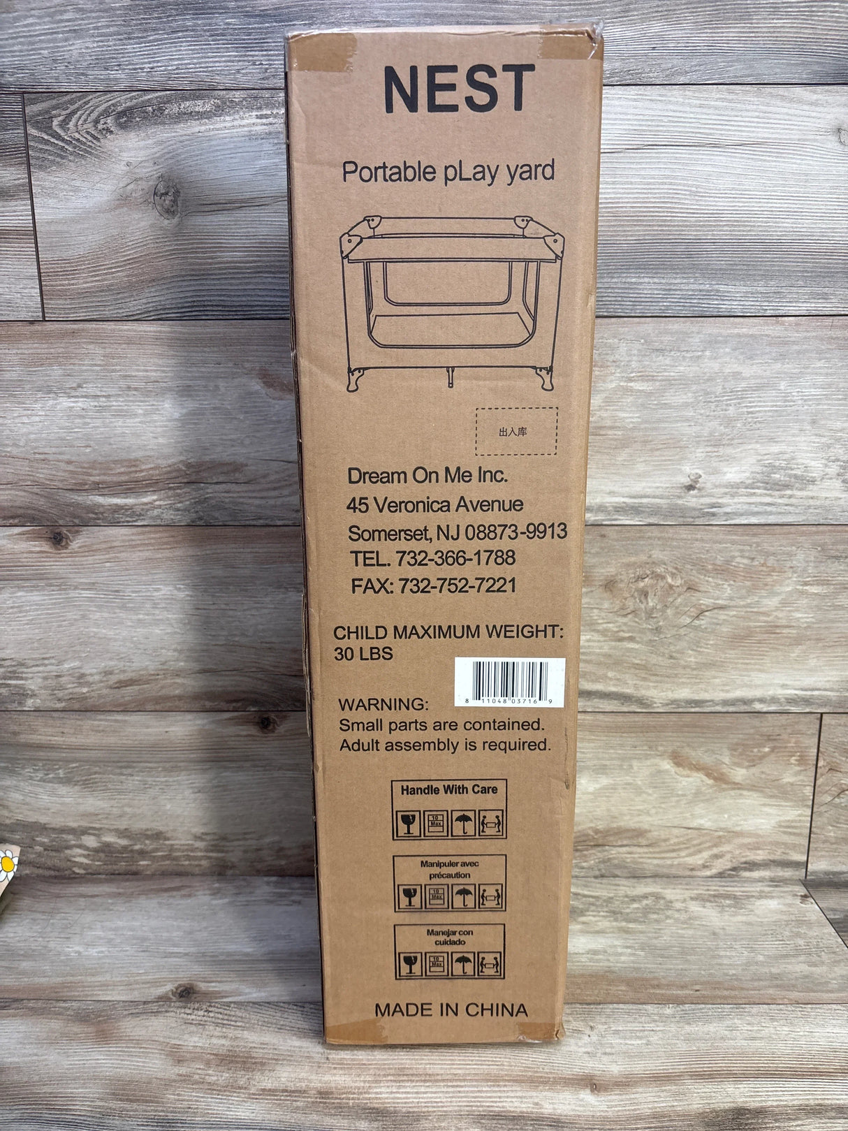 Front view of Dream On Me Nest portable playard box showing product illustration, child maximum weight 30 lbs warning and barcode on a wood floor background