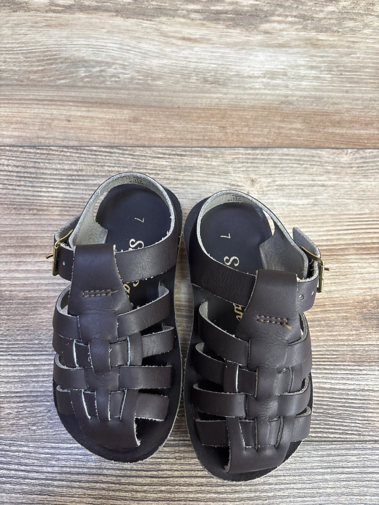 Top-down view of Salt Water Sun-San brown toddler sailor sandals showcasing woven leather upper and ankle buckle with visible size 7c on wood-look background