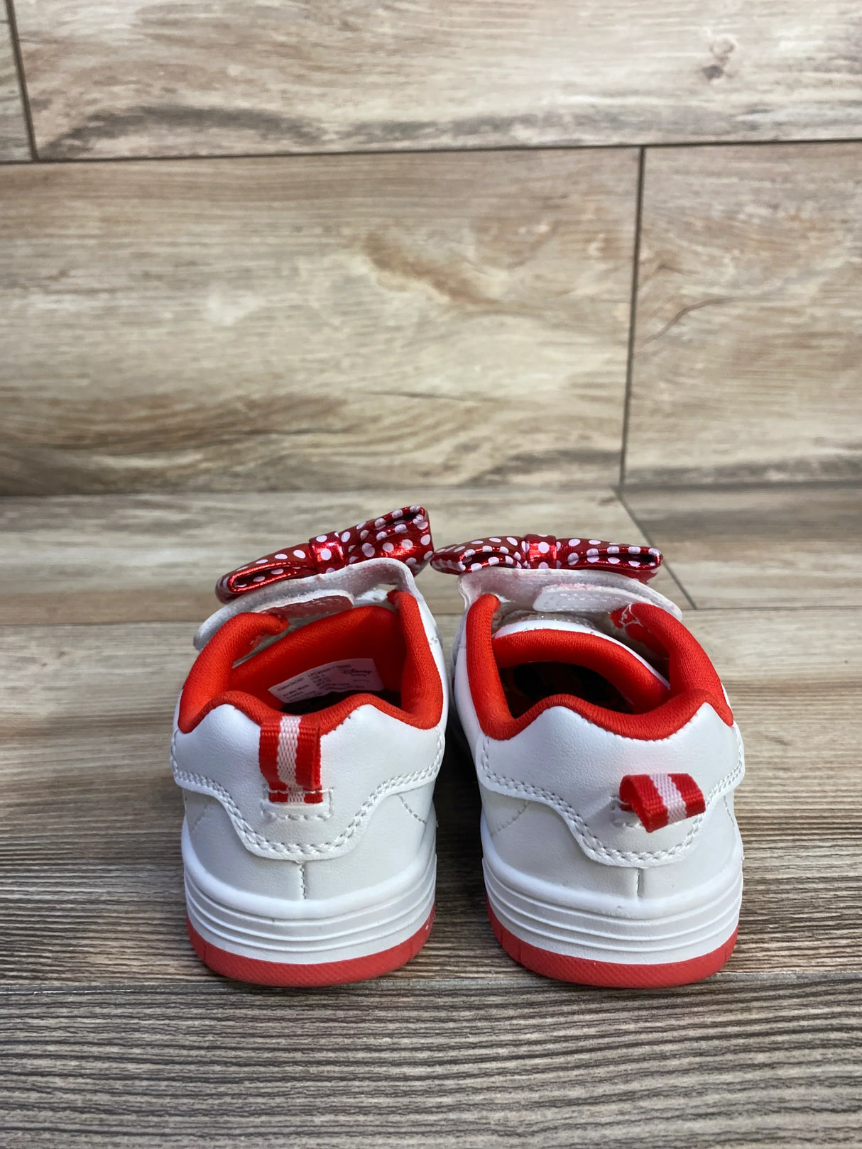 Rear view of Disney Minnie white baby sneakers with red lining, polka-dot bows and heel stripes on wood floor