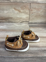 Three-quarter view of Sperry boat crib shoes in brown and tan showing moc-toe stitching, knotted laces and white soles on light wood background