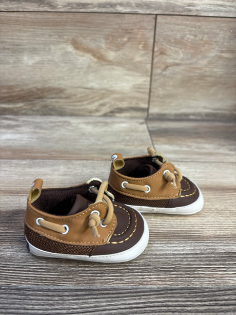 Three-quarter view of Sperry boat crib shoes in brown and tan showing moc-toe stitching, knotted laces and white soles on light wood background