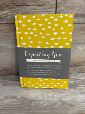 Front view of Expecting You pregnancy journal with yellow hardcover, white cloud pattern, and gray belly band on wood backdrop