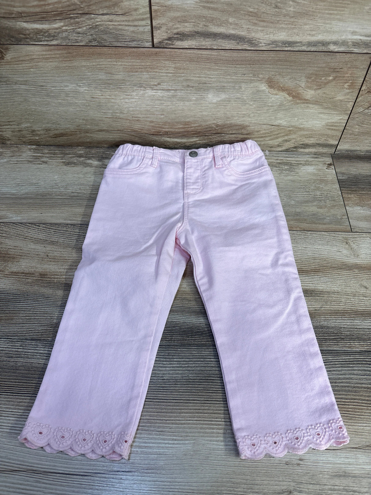 Front flat lay of pale pink OshKosh jeans with scalloped embroidered hem, elastic waistband, faux button on wood floor