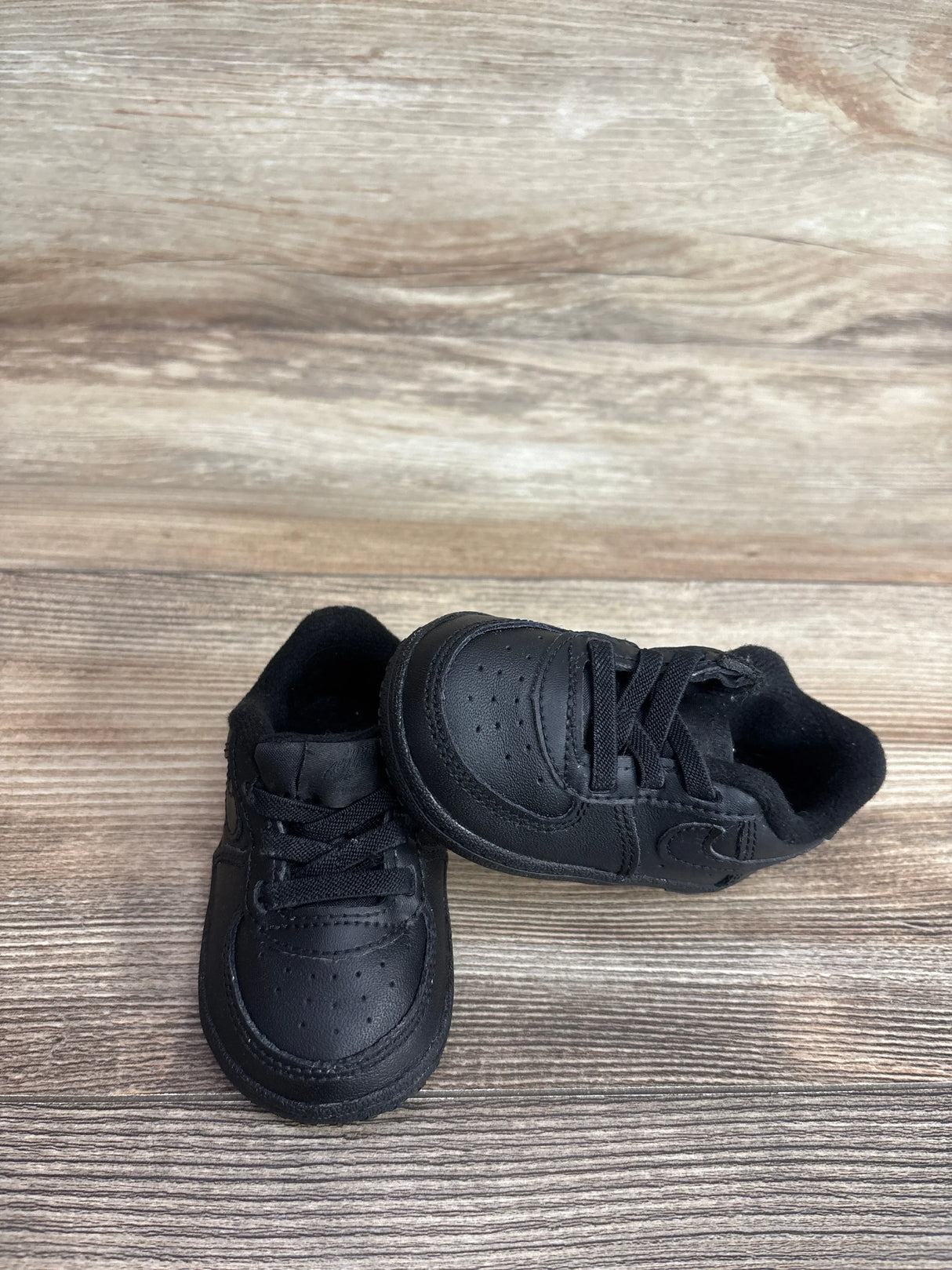 Front angle view of black Nike Air Force 1 crib shoes size 4C showcasing perforated toe, laces and Nike Swoosh with wood floor background
