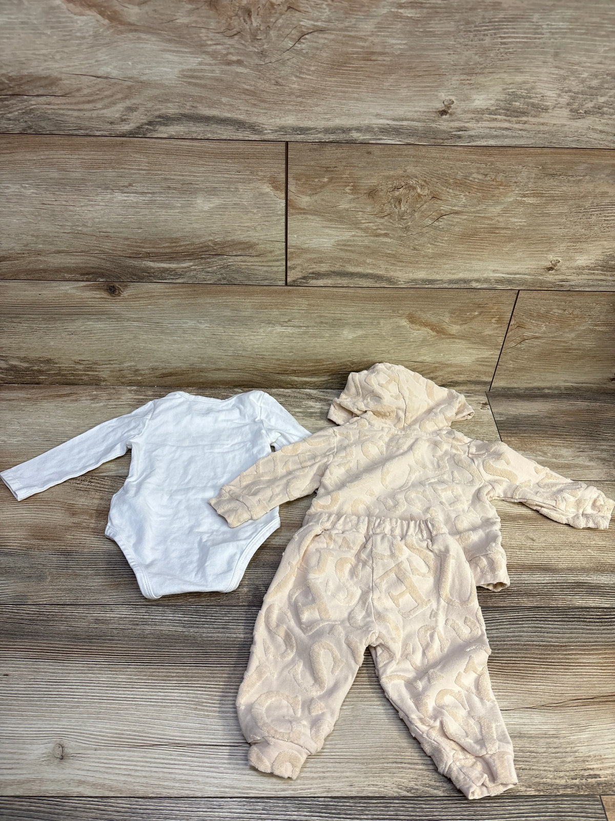 Top-down view of Guess Baby beige three-piece set with hooded patterned zip-up jacket, joggers, and white bodysuit on a wood floor