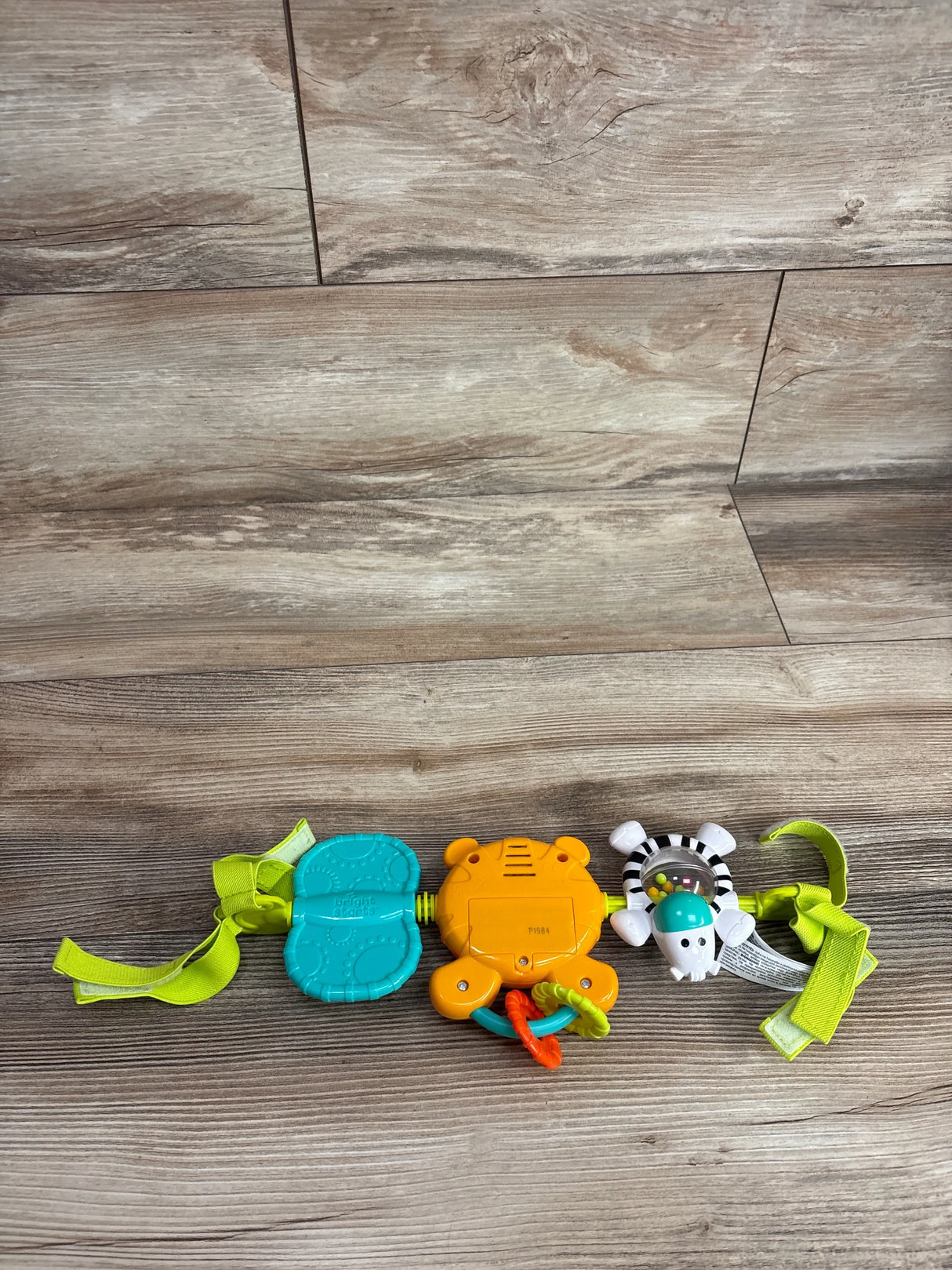 Top view of Bright Starts take along carrier toy bar showcasing teal teether orange tiger rattle and zebra clip toy with green straps on wood plank floor
