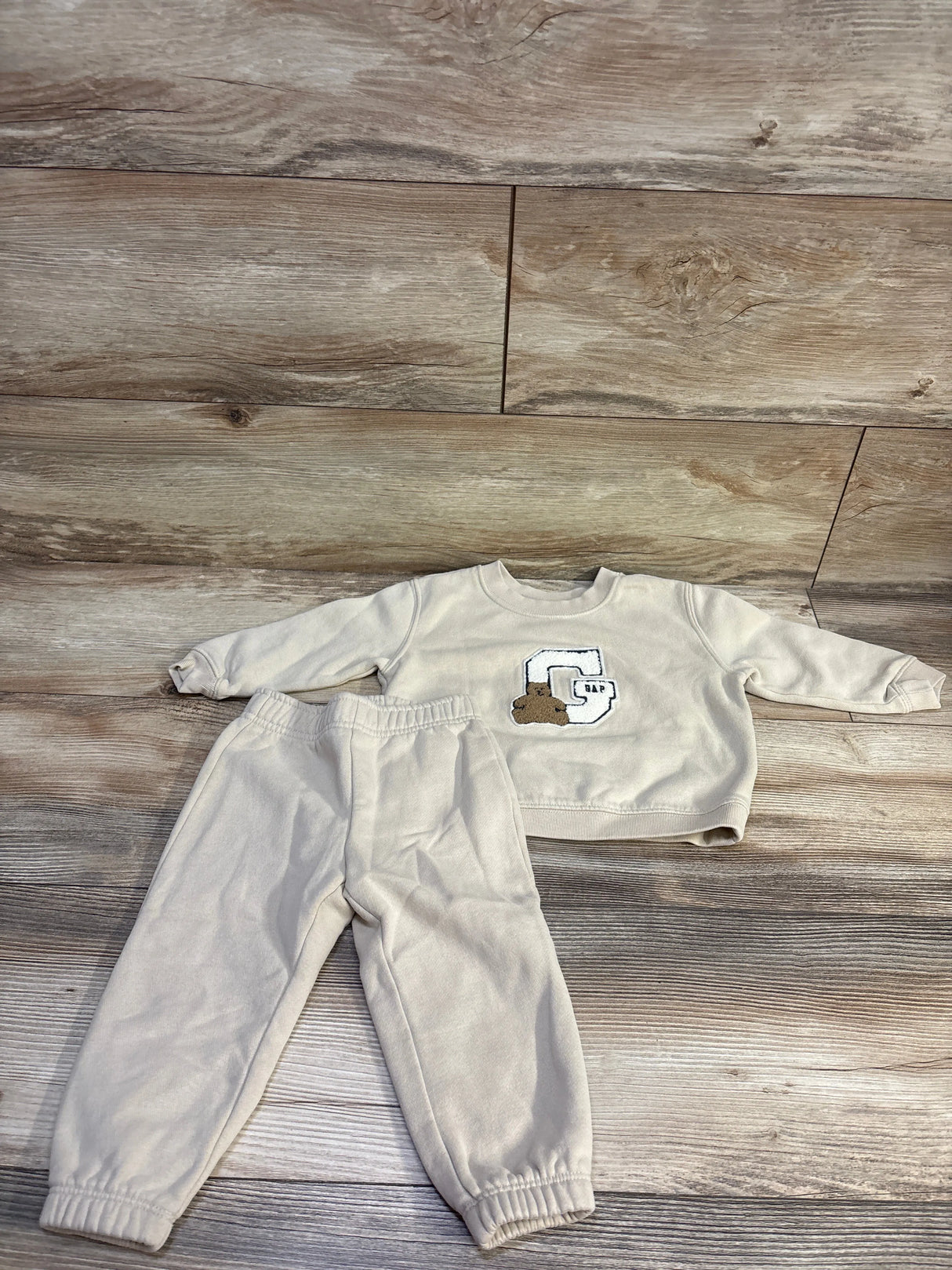 Front view of Baby Gap 2-piece pullover sweatshirt and pants set showcasing beige fabric and Gap bear logo on wood flooring