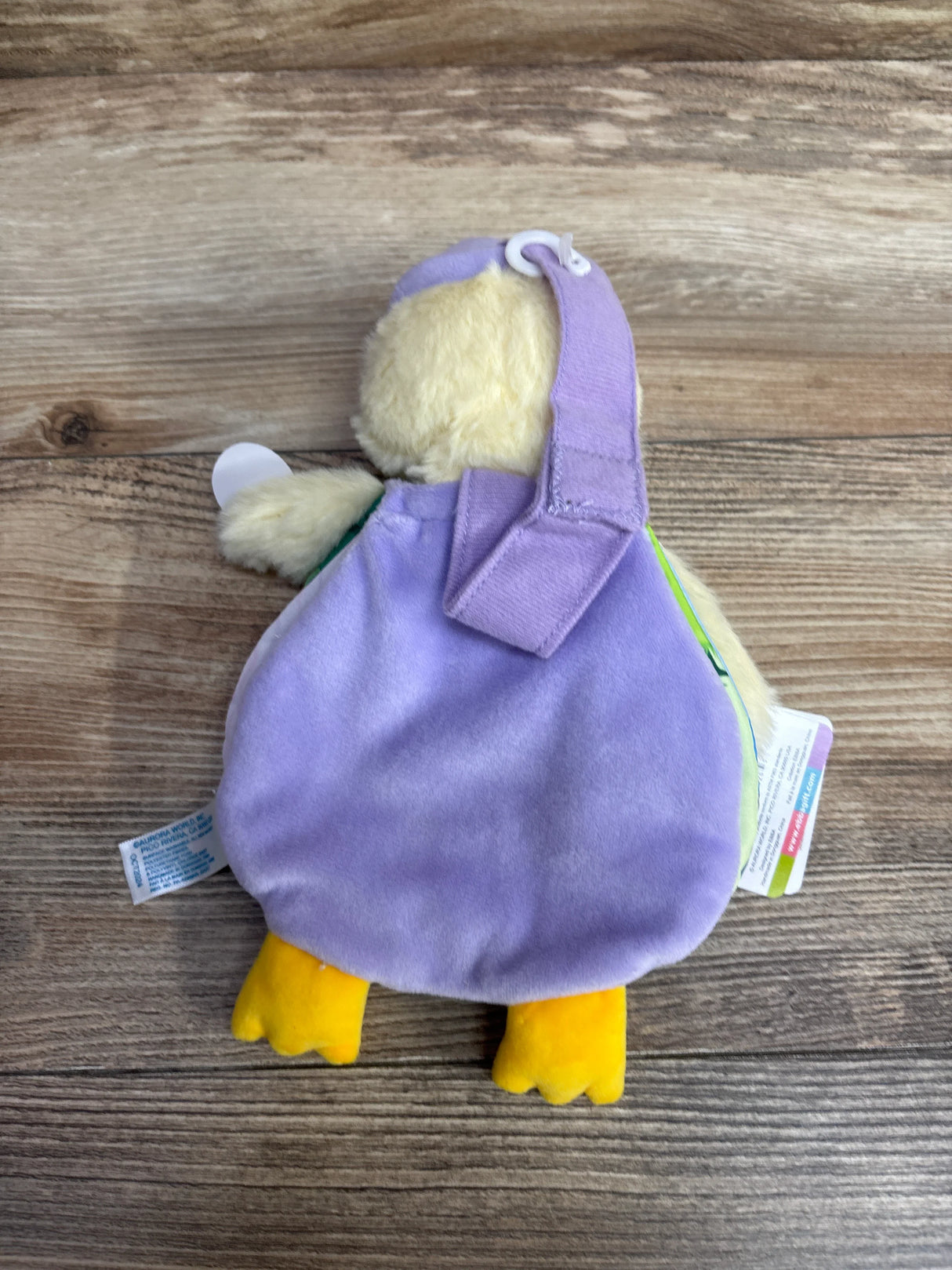 Plush yellow duck baby toy with purple fabric body and strap on wooden surface