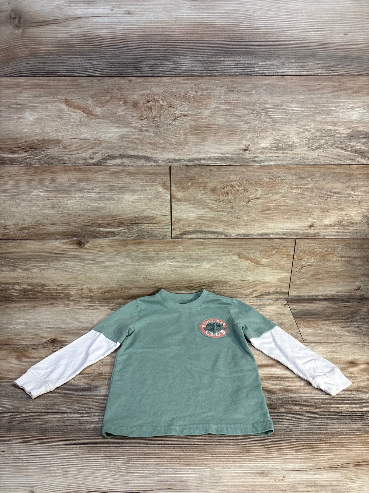 Green kids shirt with Explorer's Club logo, layered over white long sleeves, on wooden background