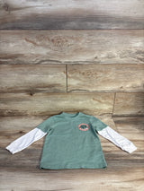 Green kids shirt with Explorer's Club logo, layered over white long sleeves, on wooden background