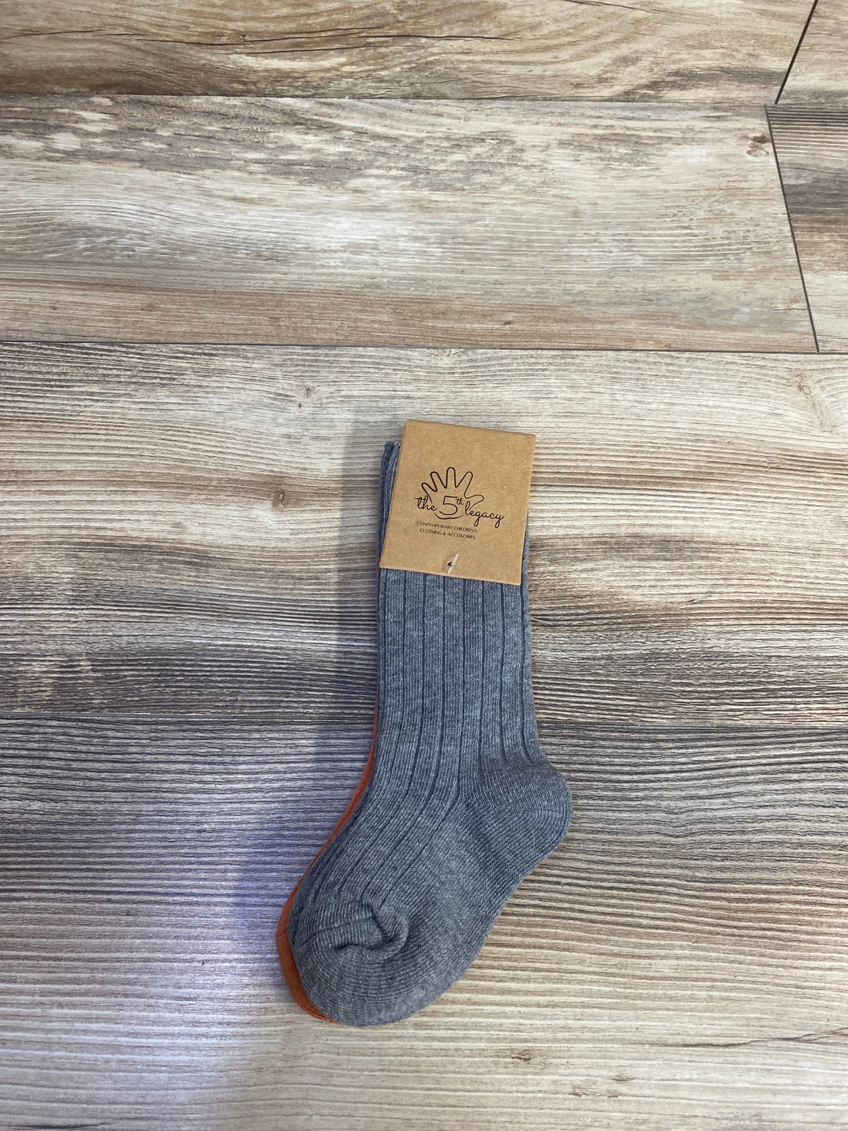 Flat lay of The 5th Legacy ribbed baby socks 2-pack size 0-12m, gray sock showing ribbed texture and orange second sock on wood floor