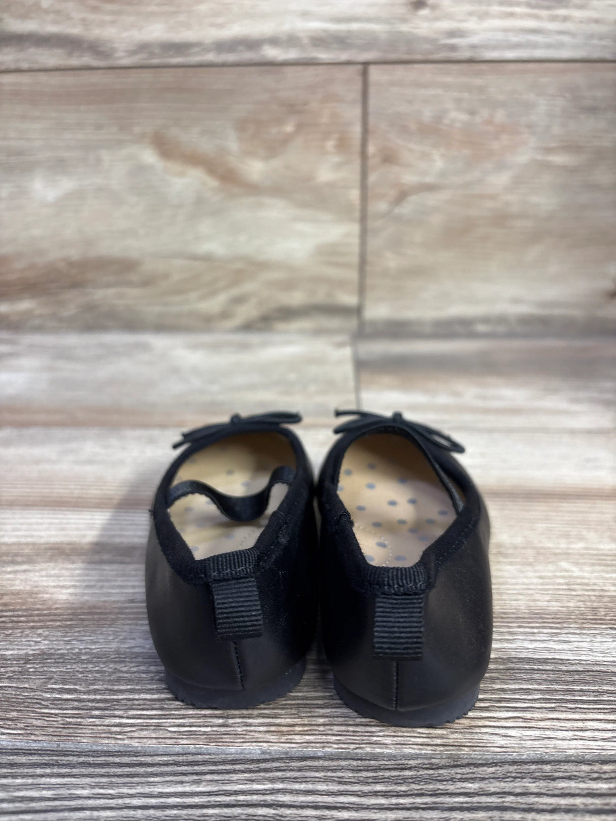 Rear view of Cat and Jack toddler black slip-on ballet flats showing elastic heel loop, bow accent, and polka-dot insole on wood floor