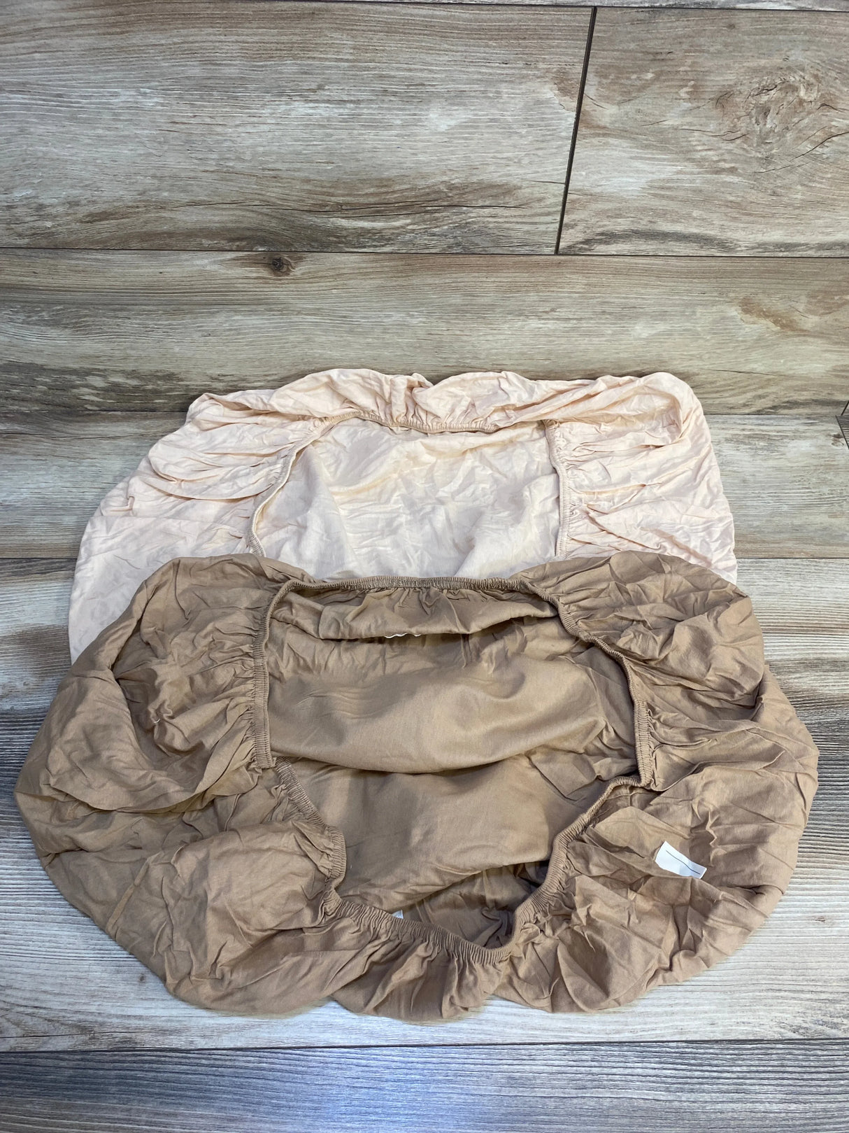 Two neutral fitted crib sheets with elastic edges on wood floor, baby bedding essentials