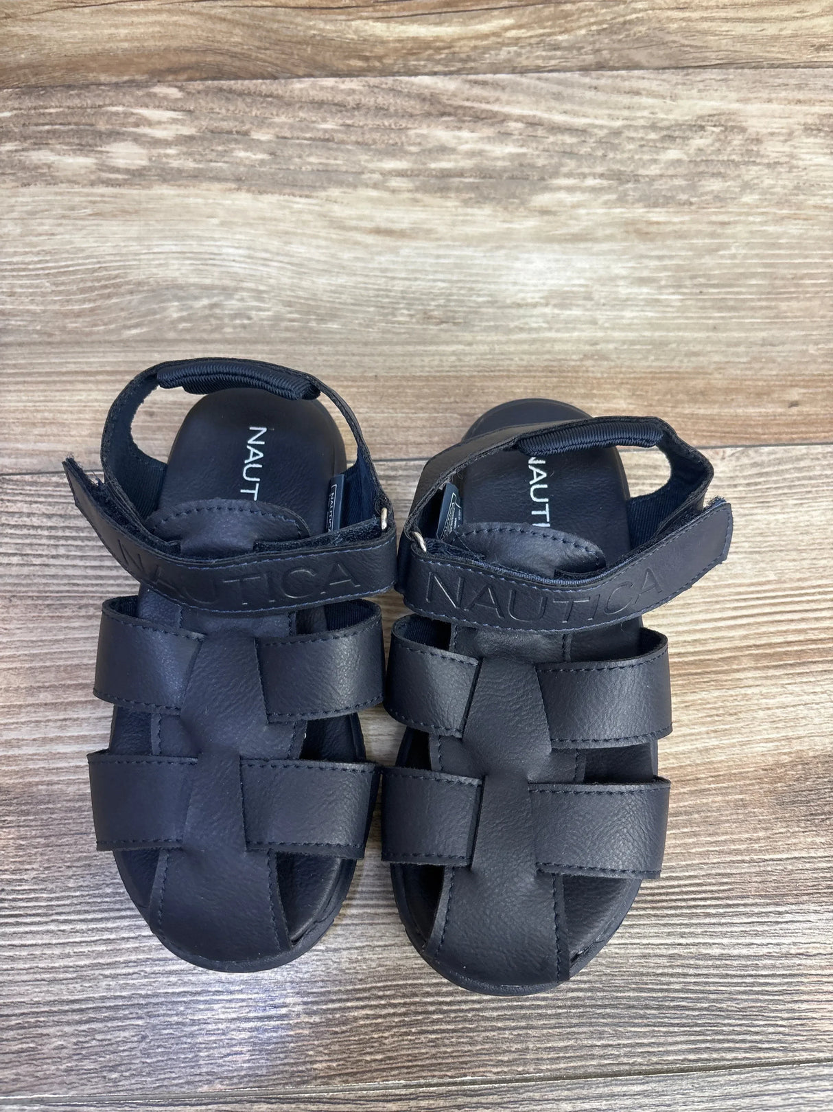 Top down view of Nautica toddler navy fisherman sandals showcasing woven straps and Velcro ankle strap with Nautica logo on wood floor