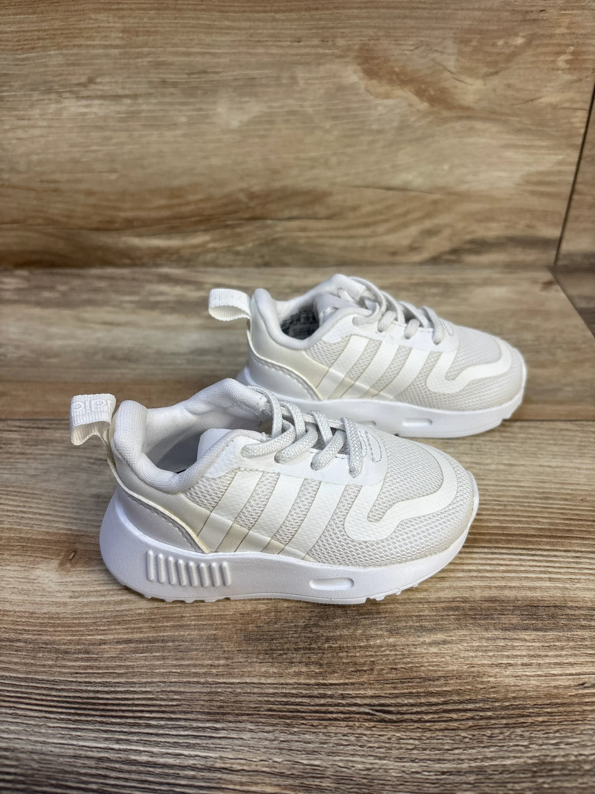 Three-quarter view of Adidas Multix toddler white sneakers showing three stripes, mesh upper and chunky sole on wood floor