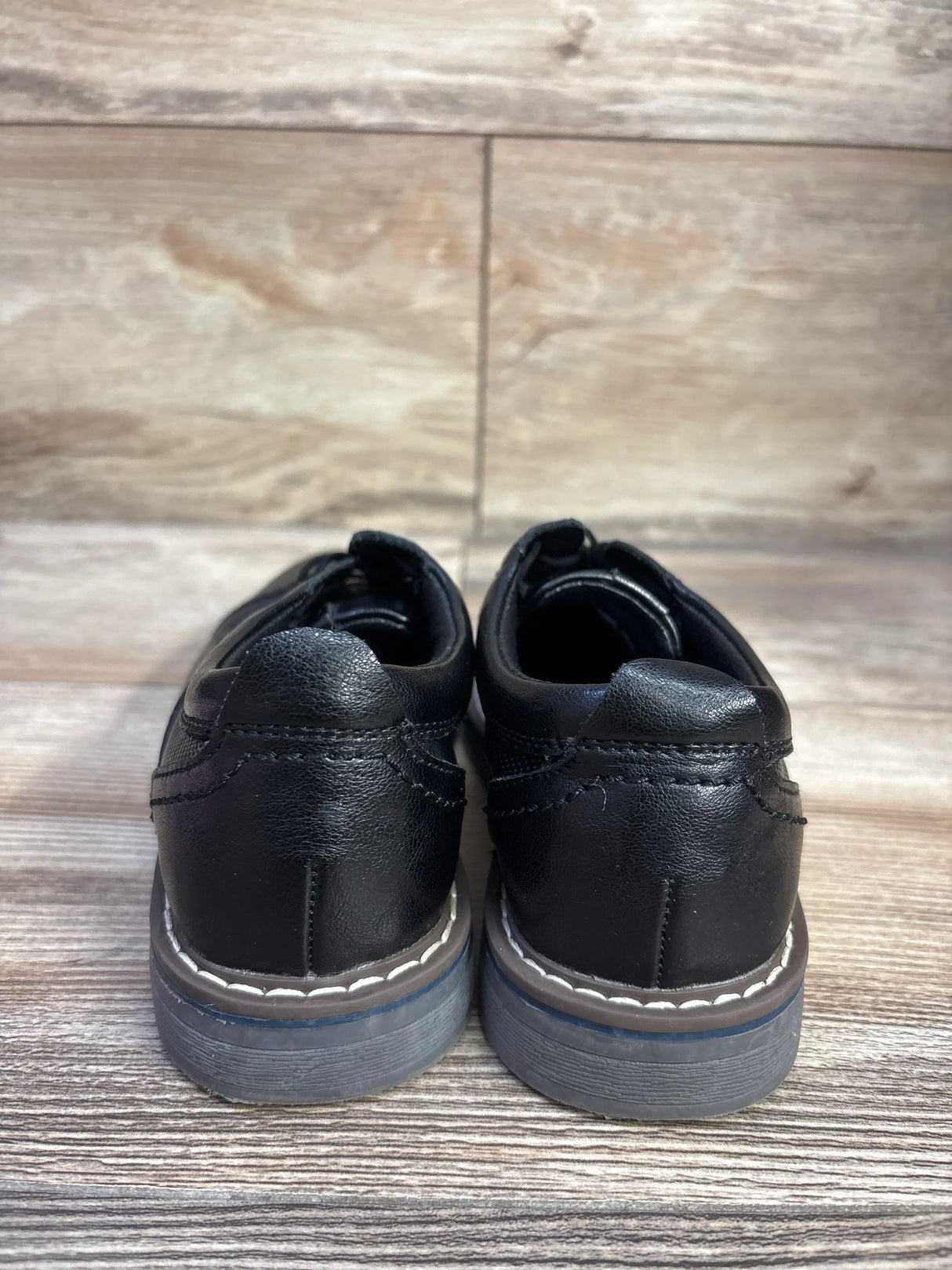 Rear view of black Steve Madden Toliverr cap toe oxford toddler shoes size 12C showing white welt stitching and gray soles on wood floor
