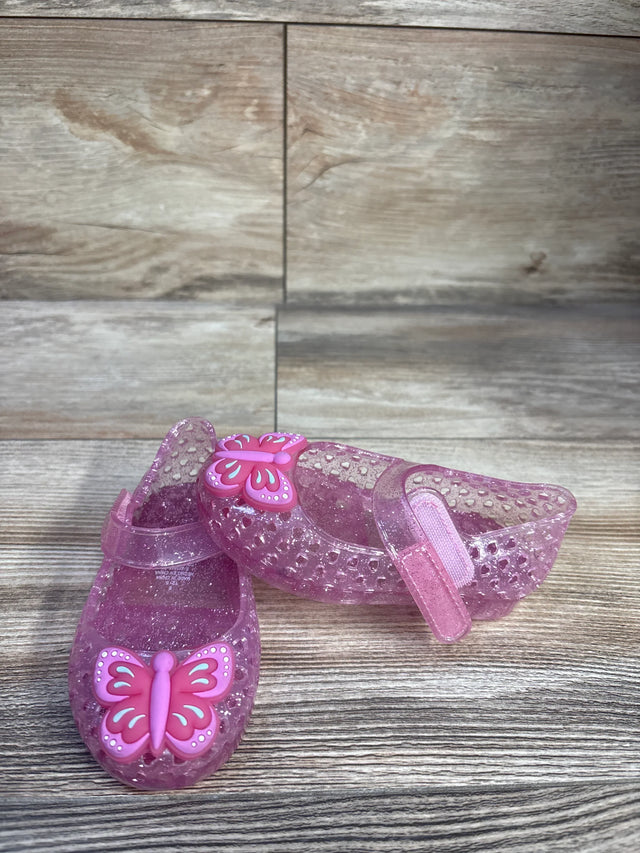 Front and side view of toddler pink glitter jelly Mary Jane flats showcasing butterfly charm and Velcro strap on wood background
