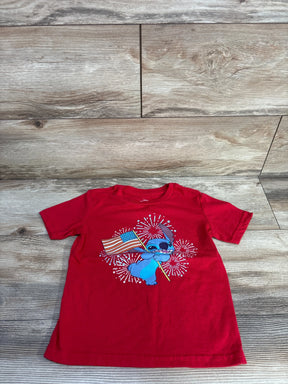 Flat-lay front view of red toddler T-shirt (5T) showing blue Stitch holding an American flag with fireworks print on a light wood floor