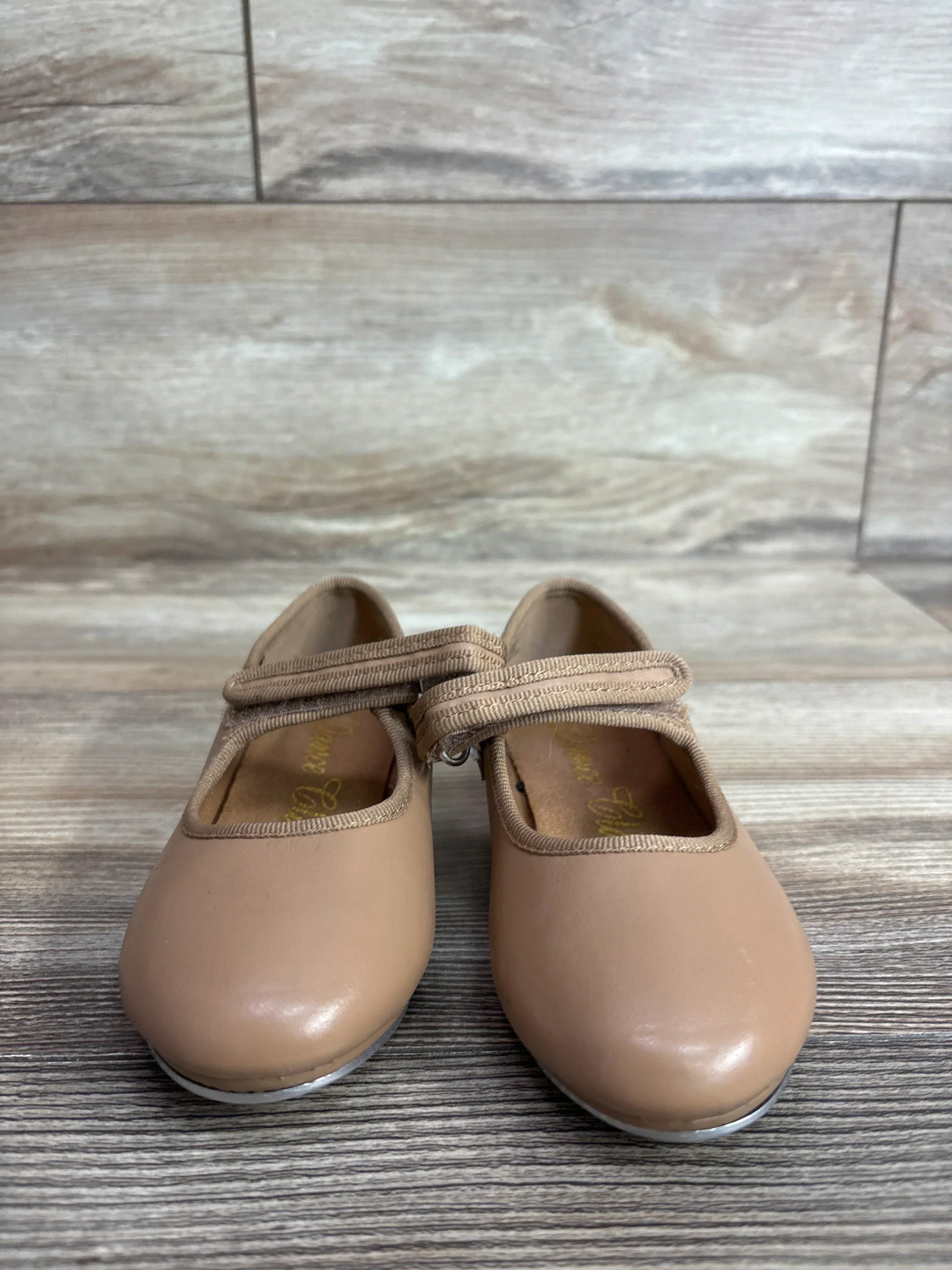 Front view of tan toddler tap shoes showcasing Mary Jane velcro strap and metal toe taps with wood plank background