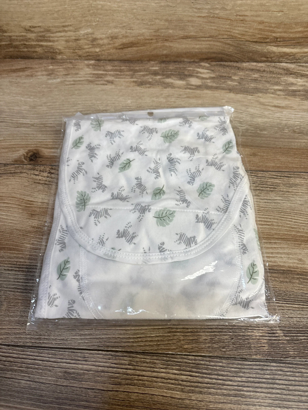 Flat lay of baby bib showcasing white fabric with zebra and green leaf print sealed in clear resale packaging on wood plank floor