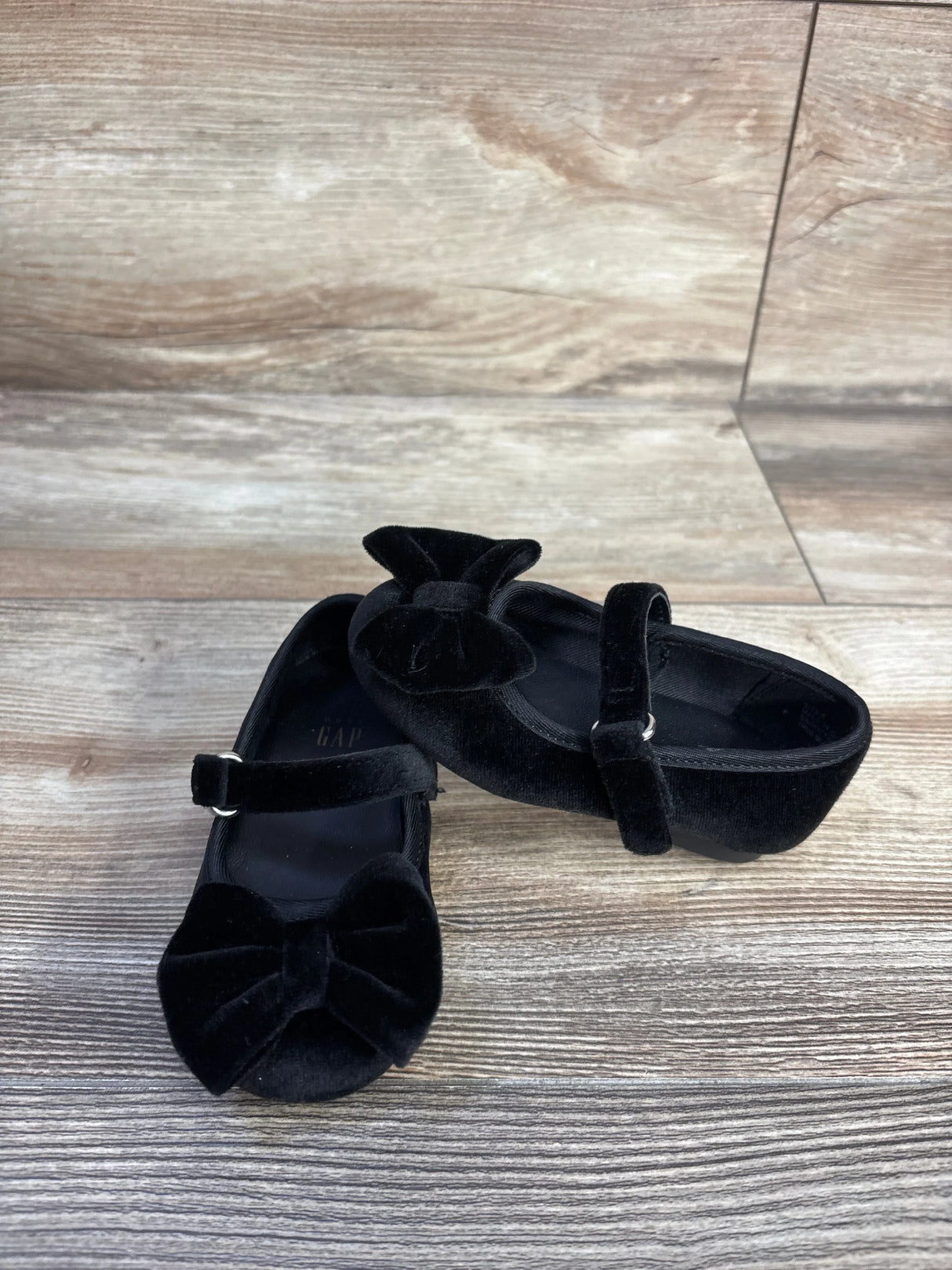 Front three-quarter view of Baby Gap velvet black Mary Jane flats showcasing bows and an adjustable strap on light wood floor