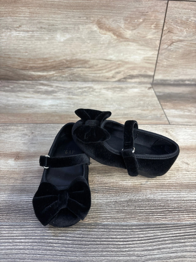 Front three-quarter view of Baby Gap velvet black Mary Jane flats showcasing bows and an adjustable strap on light wood floor