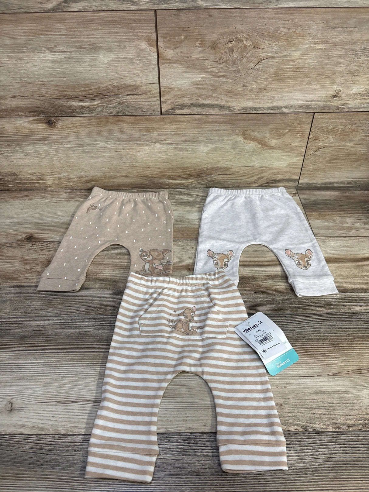 Flat lay of three Disney Baby newborn Bambi pants showing embroidered Bambi faces and tan and white stripes on wood backdrop