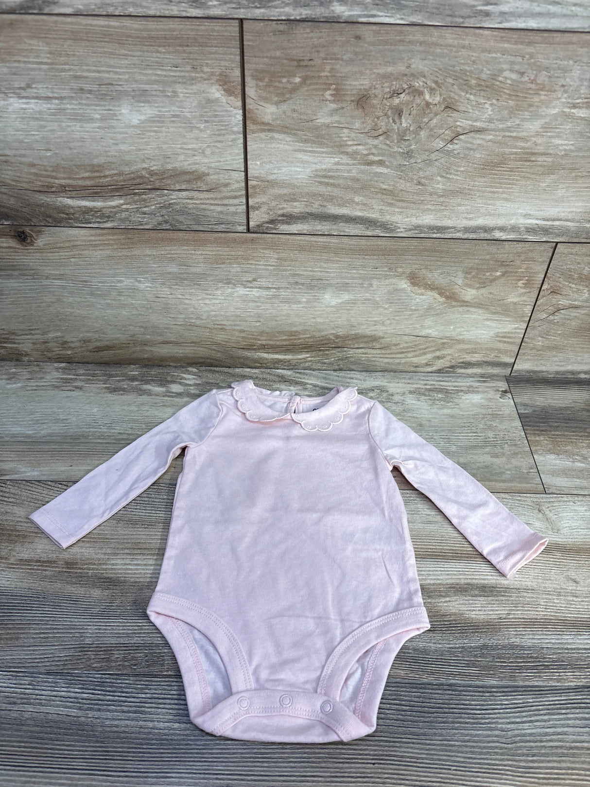 Flat lay of pink OshKosh long-sleeve baby bodysuit showing scalloped Peter Pan collar and three snap closures on wood plank background
