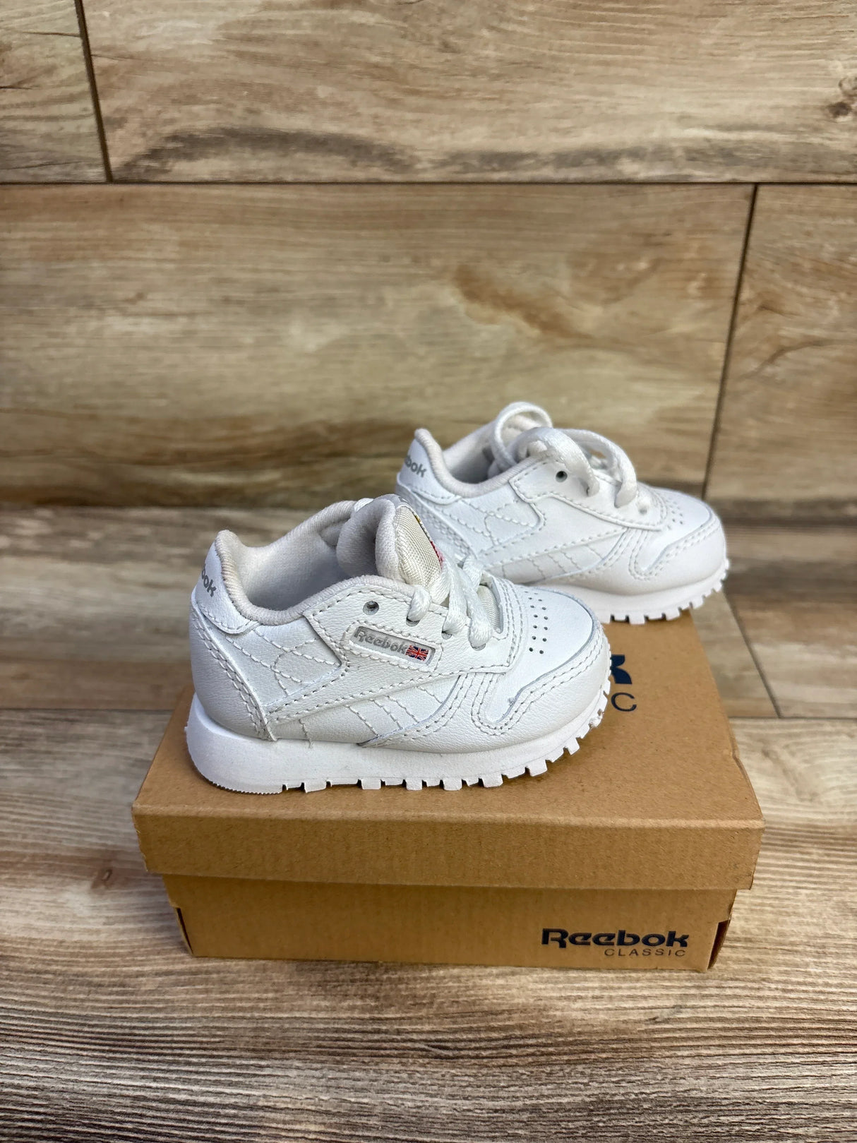 Side view of white Reebok Classic infant sneakers (size 2C) showing leather upper and tread on Reebok box, wood backdrop