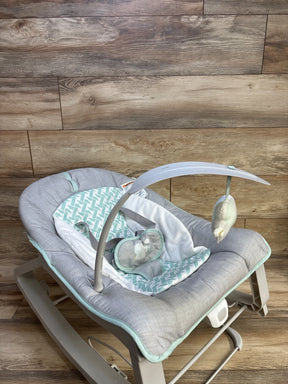 Angled view of Ingenuity Keep Cozy 3-in-1 baby bouncer showing gray and mint seat, removable toy arch, on wood floor