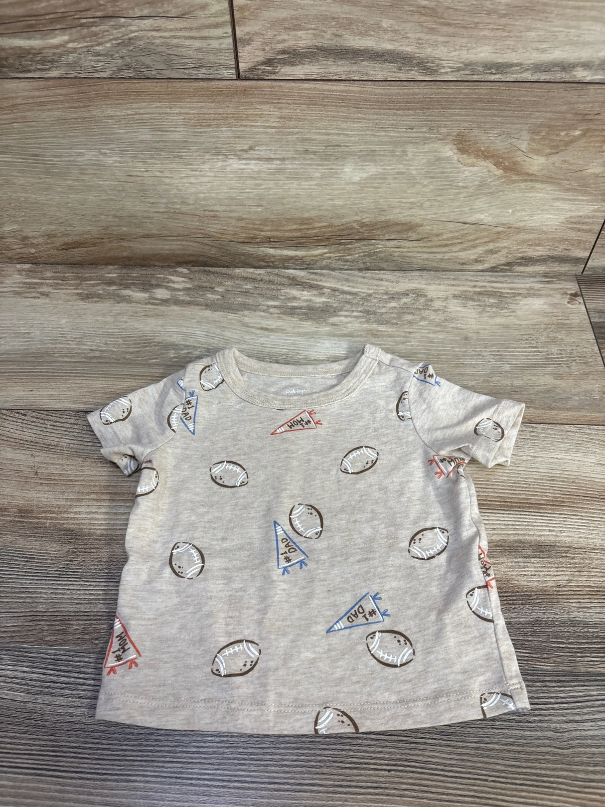 Flat lay of beige baby short-sleeve tee showing all-over football and pennant print on wood floor background