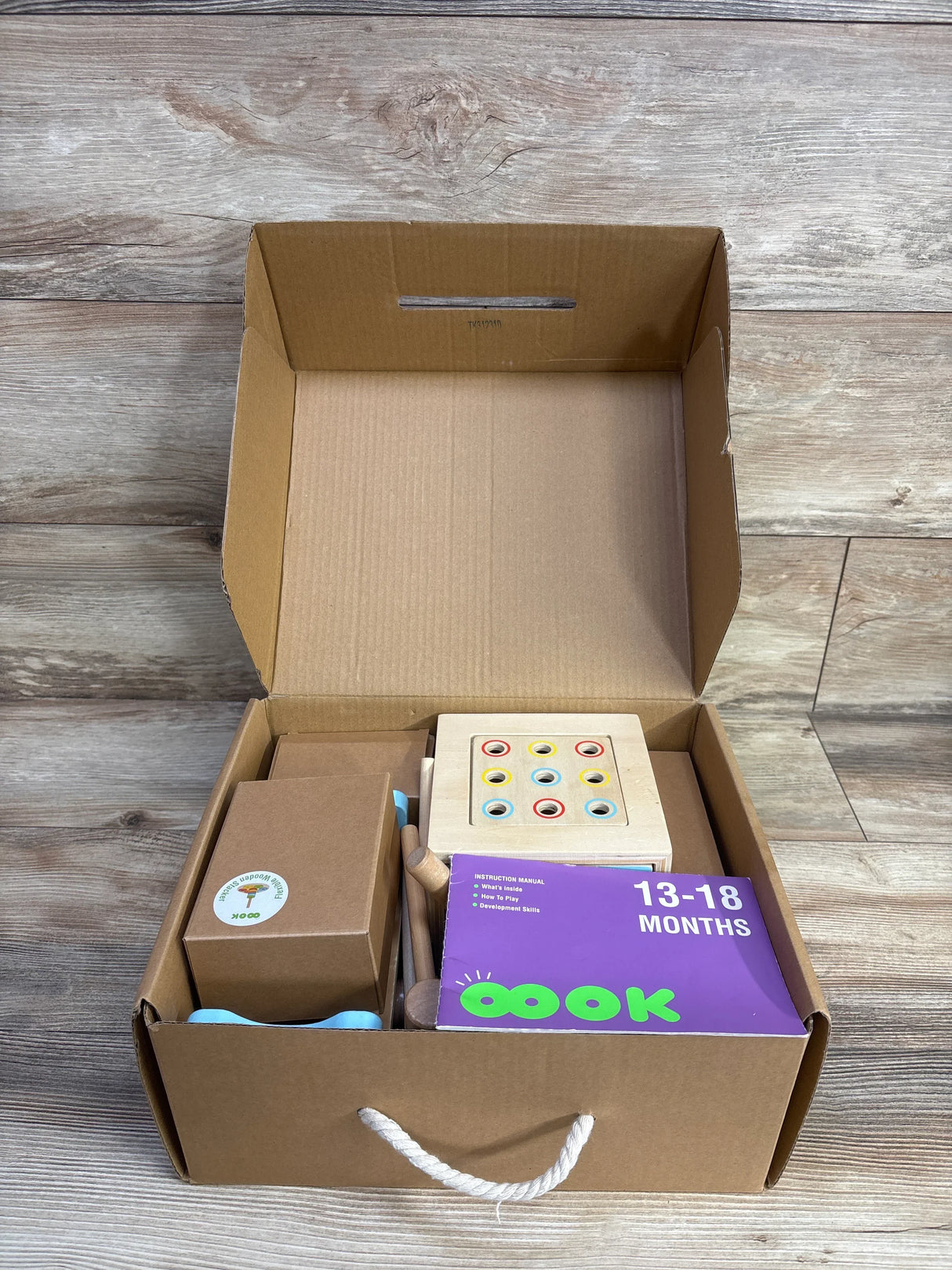 Top-down view of opened TOOKYLAND Montessori toy box showing wooden pegboard with nine colored rings, purple 13-18 months manual, and accessories in a brown box