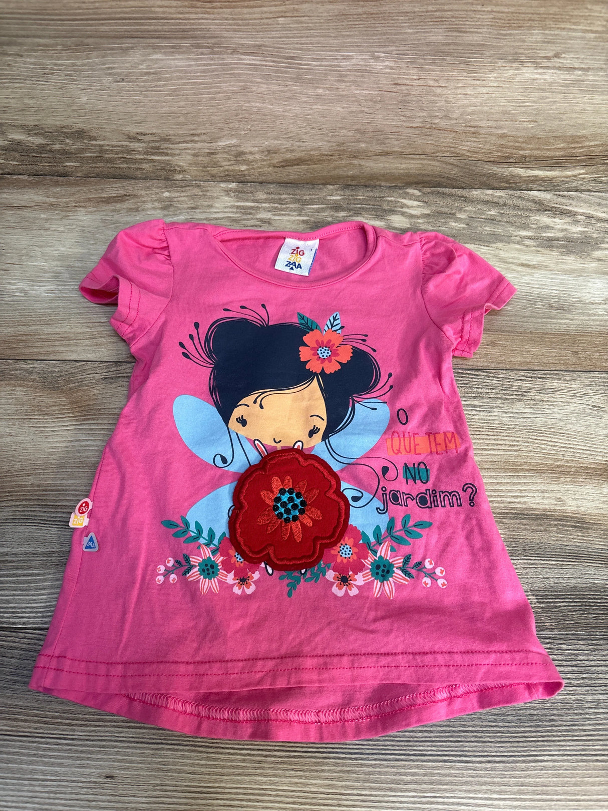 Pink kids t-shirt with fairy graphic, floral appliqué, and 'O que tem no jardim?' text, laid on wood floor