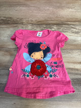 Pink kids t-shirt with fairy graphic, floral appliqué, and 'O que tem no jardim?' text, laid on wood floor