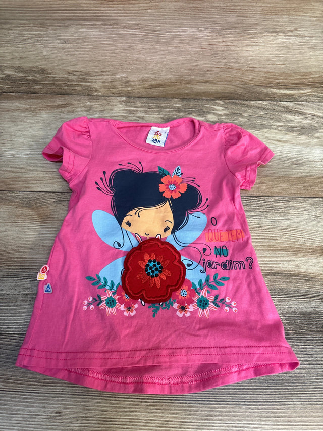 Pink kids t-shirt with fairy graphic, floral appliqué, and 'O que tem no jardim?' text, laid on wood floor