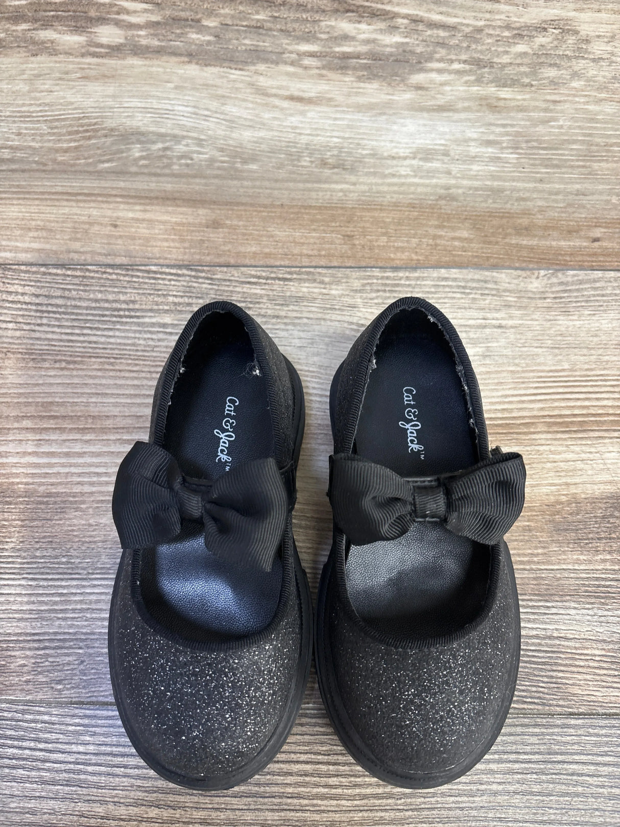 Top-down view of Cat and Jack Mary Jane flats showcasing black glitter finish, grosgrain bow strap and lug sole on wood floor