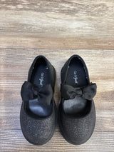 Top-down view of Cat and Jack Mary Jane flats showcasing black glitter finish, grosgrain bow strap and lug sole on wood floor