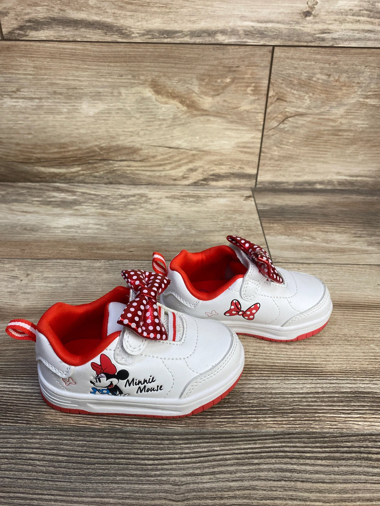 Angled view of white Minnie Mouse baby sneakers with red polka-dot bow, red lining, and Minnie graphic on wood background