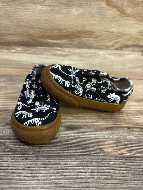 Three-quarter view of Vans Authentic dinosaur skeleton sneakers in black with gum soles on a wood-grain surface