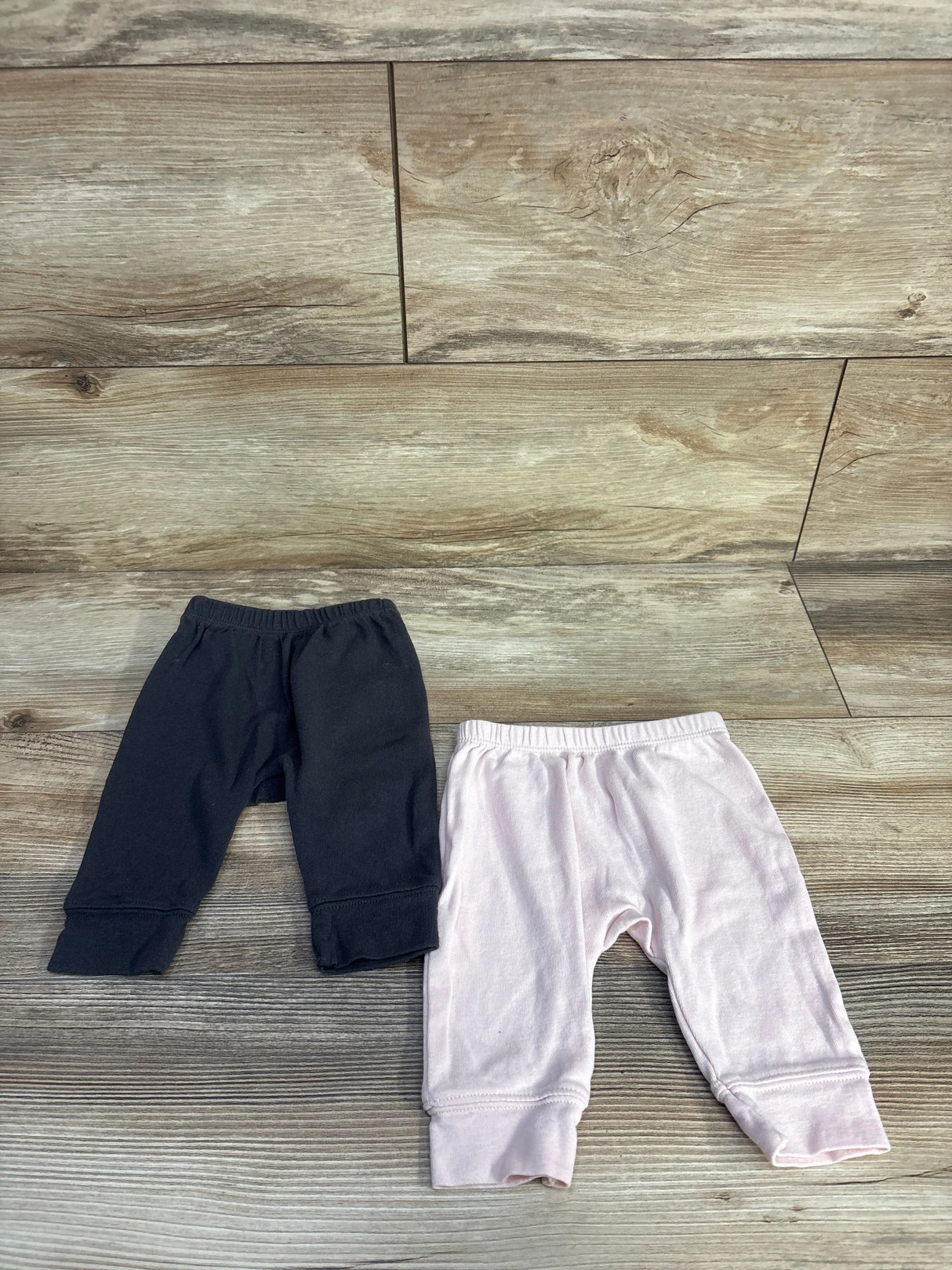 Top-down view of two Cloud Island newborn pull-on pants, grey and pale pink, showing elastic waist and cuffed ankles on wood