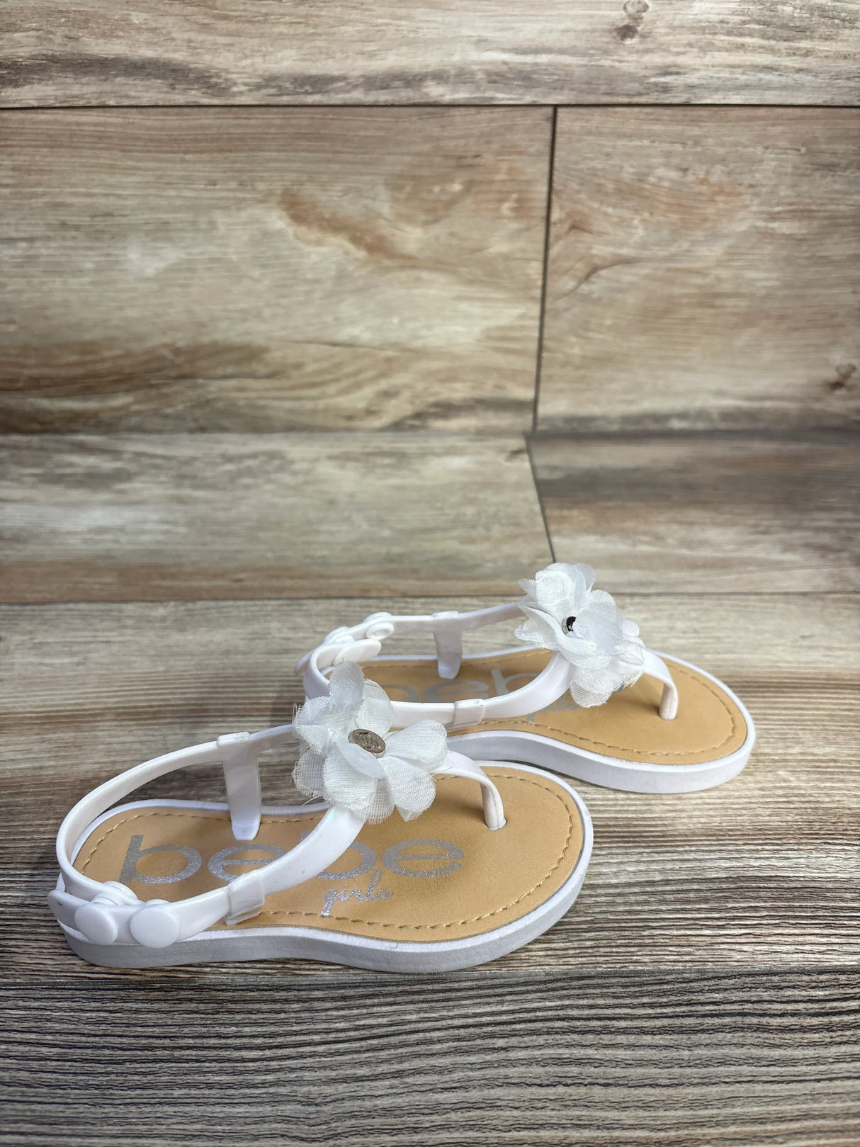 Angled view of white Bebe chiffon flower toddler sandal showcasing fabric flower embellishments and button back strap on wood backdrop